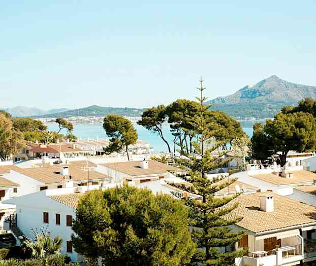 Flere hvide huse med tegltage omgivet af træer, med bjerge og hav i baggrunden. (AI-genereret tekst)