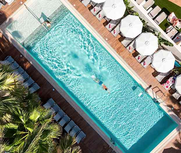 Et fugleperspektiv af en swimmingpool med en svømmende person og liggestole med parasoller omkring. (AI-genereret tekst)