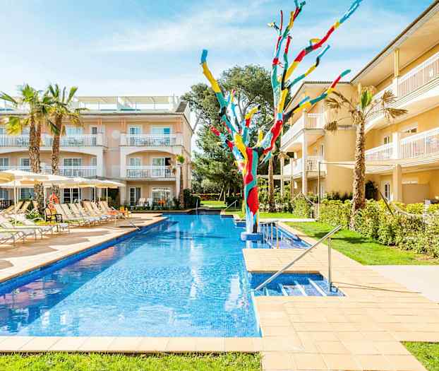 Udendørsområde med swimmingpool, liggestole og parasoller omgivet af bygninger og to farverige kunstværker. (AI-genereret tekst)