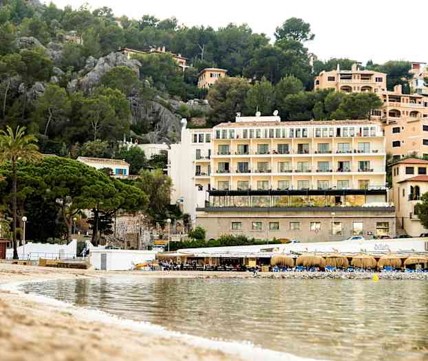Billede af en strand med klart vand, en lille restaurantbygning og bygninger med flere etager omgivet af grønt i baggrunden. (AI-genereret tekst)