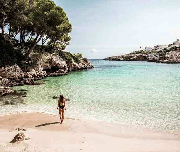 Kvinde står på en sandstrand ved en bugt med klart vand omgivet af klipper og grønne områder. (AI-genereret tekst)