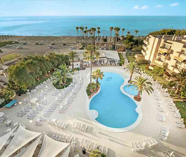 Luftfoto af hotelområde med pool, liggestole og palmer, samt nærhed til stranden. (AI-genereret tekst)