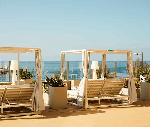 To liggestole med hvid tagstruktur og gardiner på en terrasse med udsigt over havet. (AI-genereret tekst)