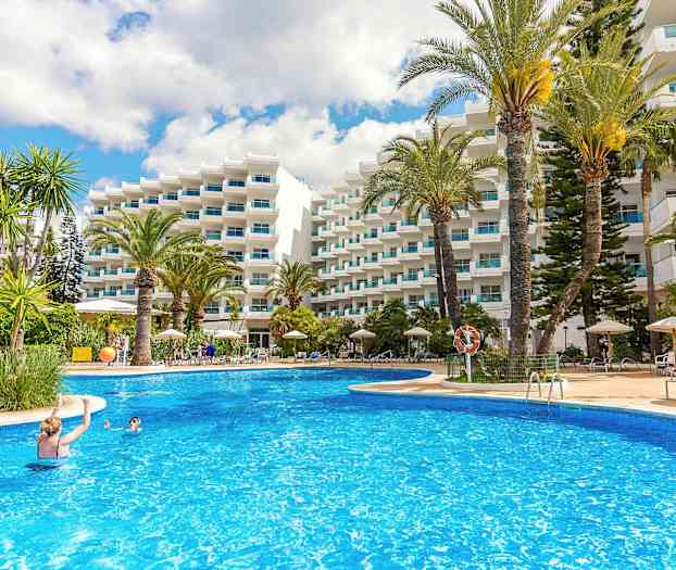 En udendørs swimmingpool omgivet af liggestole, parasoller og høje palmer, foran en hvid bygning med altaner. (AI-genereret tekst)