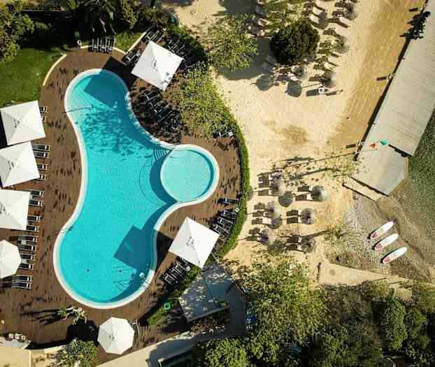 Luftfoto af en pool omgivet af liggestole og parasoller, med en tilstødende strand og bro. (AI-genereret tekst)