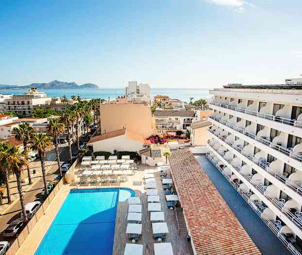 Udsigt fra oven af en hotelbygning, pool og omkringliggende område med havet i baggrunden. (AI-genereret tekst)