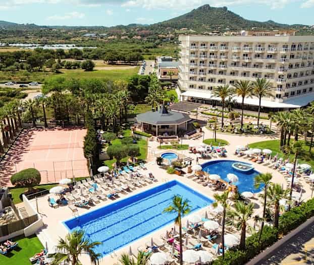 Hotelkompleks med swimmingpools, liggestole, tennisbane og have i forgrunden samt omkringliggende grønne områder og bjerge i baggrunden. (AI-genereret tekst)