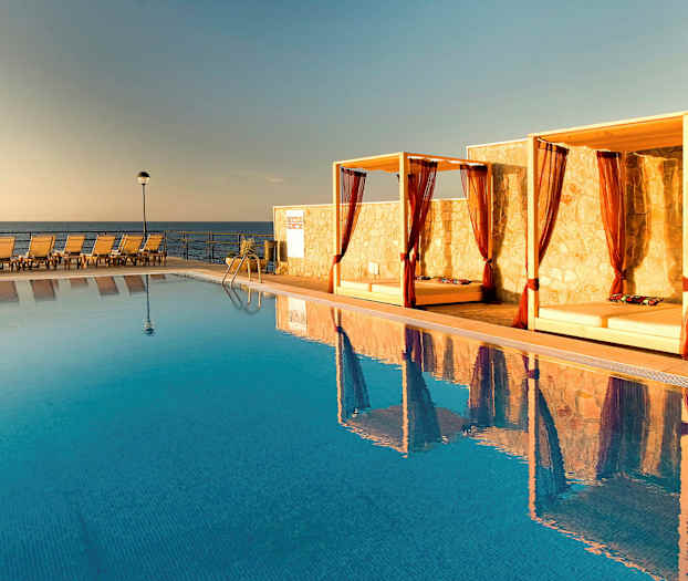 Billede af en pool med liggestole og baldakiner ved kanten samt havet i baggrunden. (AI-genereret tekst)