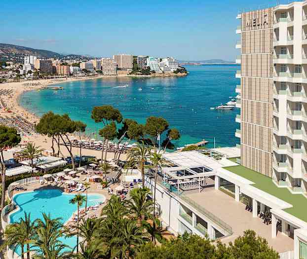 En strand med klart blåt vand, omgivet af hotelbygninger, træer og en swimmingpool. (AI-genereret tekst)