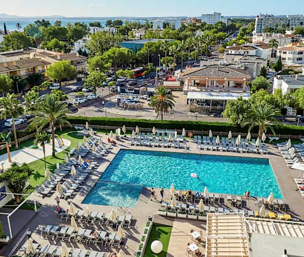 En pool omgivet af liggestole, med omkringliggende grønne områder og bygninger i baggrunden. (AI-genereret tekst)