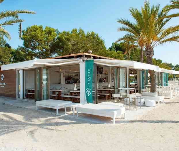 En udendørs lounge og restaurant ved stranden omgivet af palmer. (AI-genereret tekst)