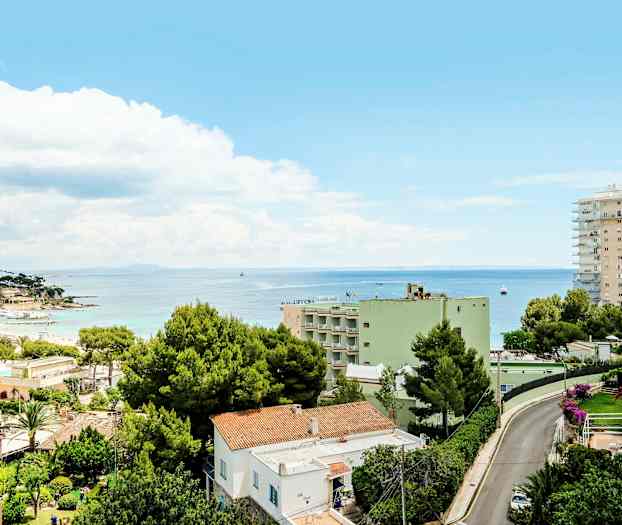 Udsigt over en by med boliger og grønne træer nær kysten. I baggrunden ses havet under en blå himmel med skyer. (AI-genereret tekst)