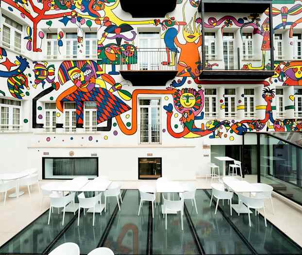 En gårdhave med hvide borde og stole samt en farverig vægmaleri på husfacaden. (AI-genereret tekst)