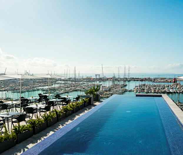 En infinitypool på en terrasse med udsigt over en marina fyldt med både og havet i baggrunden. Der er siddepladser under parasoller ved siden af poolen. (AI-genereret tekst)