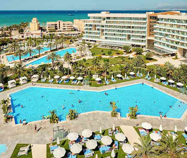 Udsigt over et feriested med swimmingpools, liggestole og omkringliggende hotelbygninger samt havet i baggrunden. (AI-genereret tekst)