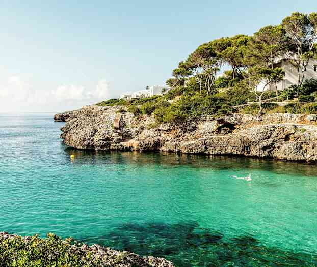 En kystlinje med klipper og klart turkis vand, et hus er synligt blandt træerne. (AI-genereret tekst)