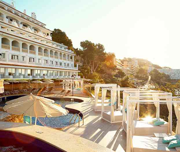 Hotellets facade i hvidt med arkader, swimmingpool og liggestole med parasoller og dagsenge, omgivet af træer og bygninger. (AI-genereret tekst)