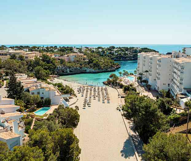 En strand med parasoller omgivet af bygninger og vegetation, med havet i baggrunden. (AI-genereret tekst)