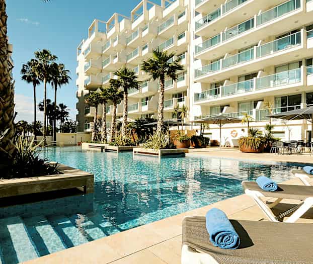 Billede af en hotelpool med solsenge og palmer omgivet af et moderne bygningskompleks. (AI-genereret tekst)