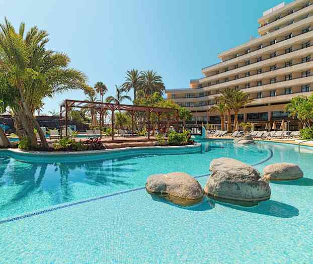 Udendørs pool med klart vand, omgivet af palmer og solsenge foran en hotelbygning. (AI-genereret tekst)