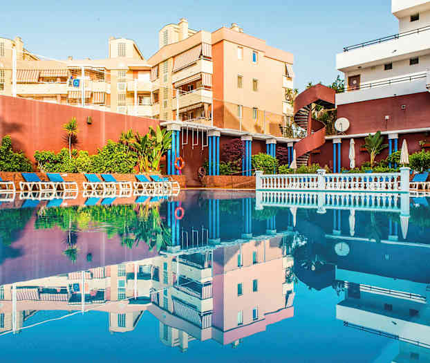 En udendørs pool omgivet af liggestole og grønne planter, med bygninger i baggrunden. (AI-genereret tekst)