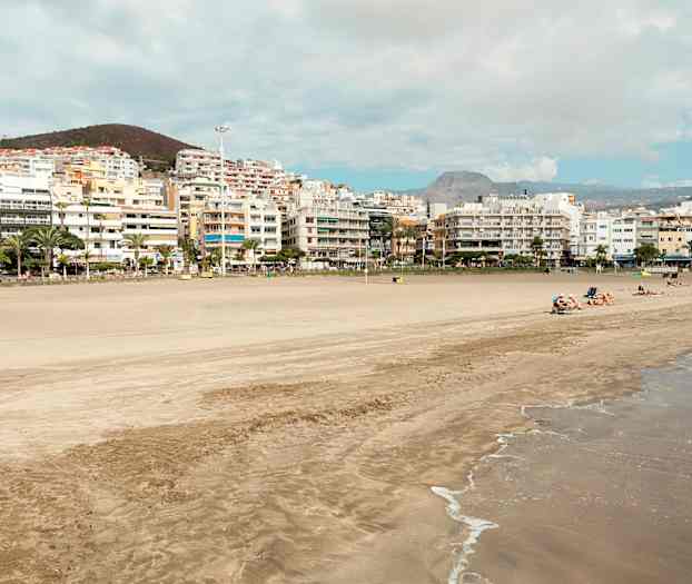 Strand med sand og bølger, omgivet af bygninger og bakker i baggrunden. (AI-genereret tekst)