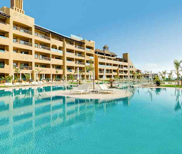 Hotellets udendørs pool med liggestole og omkringliggende bygninger. (AI-genereret tekst)