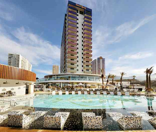 Et hotel med en stor pool og omkringliggende bygninger under en blå himmel. (AI-genereret tekst)