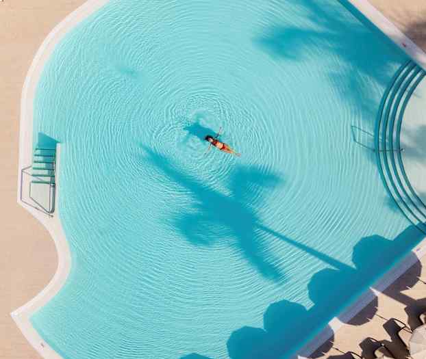 En kvinde flyder i en hotelpool, omgivet af parasoller og palmer. (AI-genereret tekst)