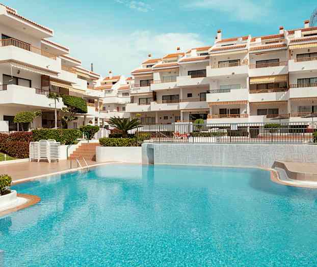 Hvid bygning med terrasser og en turkis pool i forgrunden. (AI-genereret tekst)