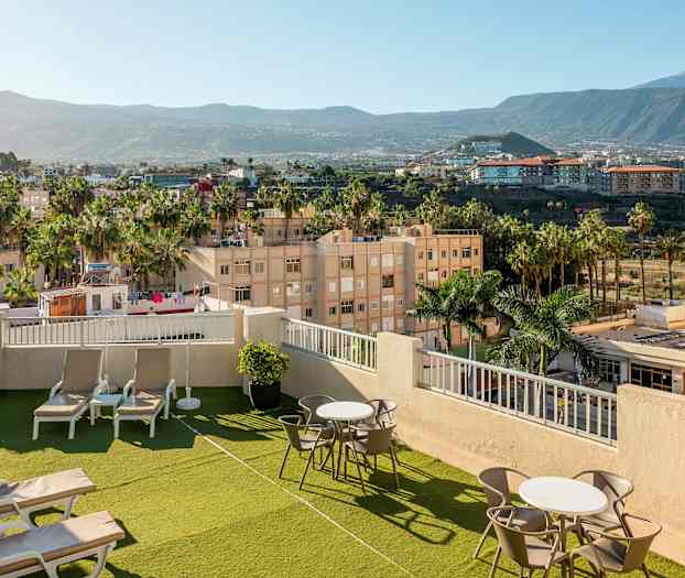 Billede af en hoteltagterrasse med solsenge og borde, omgivet af grønne områder og palmer, med udsigt over nærliggende byområde og bjerge. (AI-genereret tekst)