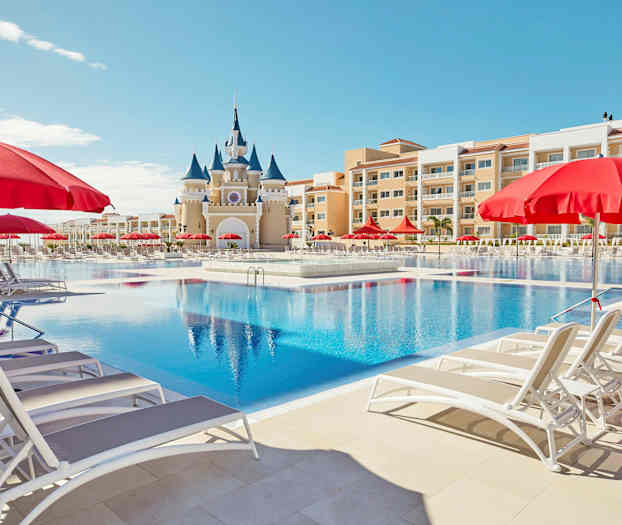 En stor swimmingpool med liggestole og røde parasoller samt en bygning med et slotslignende udseende i baggrunden. (AI-genereret tekst)