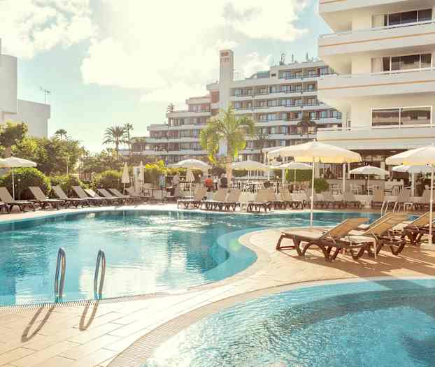 Hotelpool med liggestole og parasoller, omgivet af bygninger. (AI-genereret tekst)