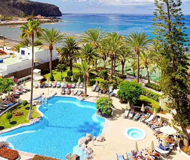 Hotelpool med liggestole og parasoller, omgivet af tropiske palmer og kystlandskab. (AI-genereret tekst)