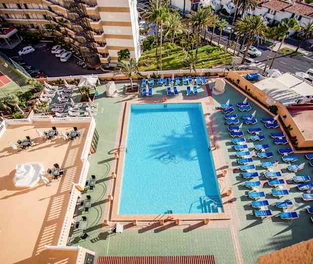 Udsigt over en pool og liggestole ved et hotel, med udsigt til omkringliggende bygninger og grønne områder. (AI-genereret tekst)