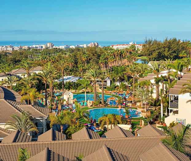 Udsigt over et feriehotel med palmer, swimmingpools og bygninger, med havet og en by i baggrunden. (AI-genereret tekst)