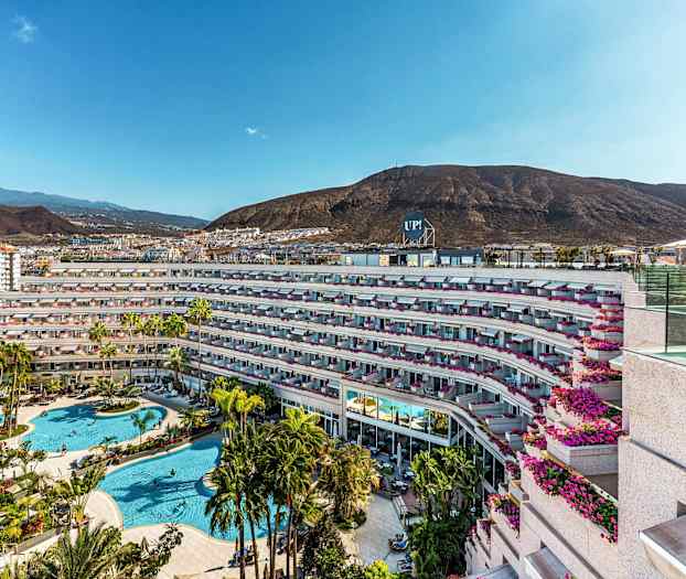 Billede af hotelkompleks med poolområde og omkringliggende bjerge. (AI-genereret tekst)