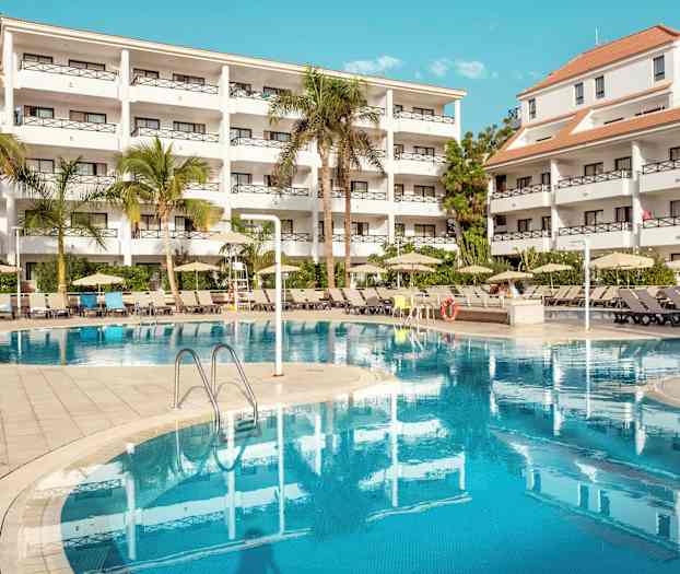 Billede af et hotelkompleks med hvide bygninger, træer og en pool. (AI-genereret tekst)