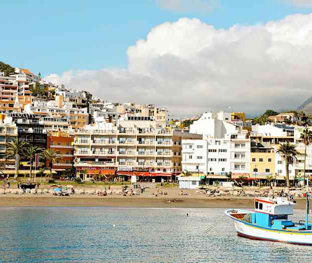 En strand med liggestole, en fiskerbåd på vandet og bygninger bagved. (AI-genereret tekst)