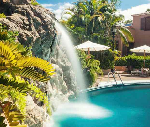 Vandfald, der strømmer ned i en swimmingpool, omgivet af grønne planter og liggestole. (AI-genereret tekst)