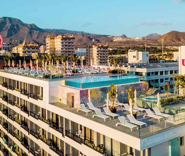 Hotelbygning med en infinity-pool og solsenge på tagterrassen, i baggrunden bjerge og byer. (AI-genereret tekst)