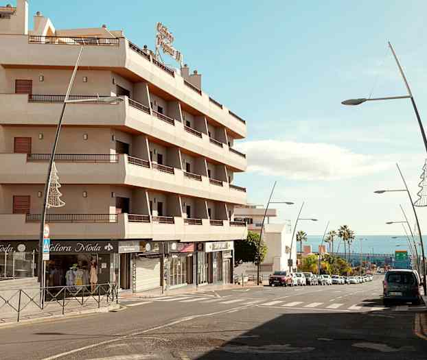 En bygning med altaner ved en gade med biler og gadelamper, med havet i baggrunden. (AI-genereret tekst)