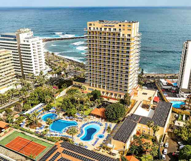 Hotelanlæg med poolområde og tennisbane, ved havet, omgivet af høje bygninger. (AI-genereret tekst)