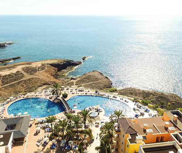 Luftfoto af et resort med swimmingpool, liggestole og havudsigt. (AI-genereret tekst)