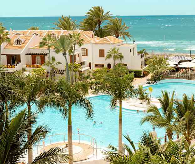 Hotelbygninger med terracottafarvede tage ved en pool omgivet af palmer og havet i baggrunden. (AI-genereret tekst)