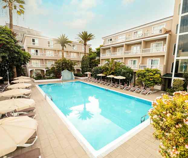 Hotelområde med en udendørs pool omgivet af liggestole og parasoller samt bygninger med altaner. (AI-genereret tekst)