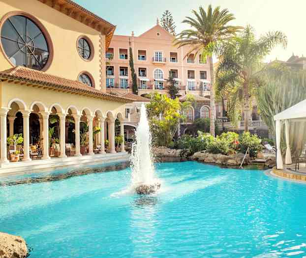 Hotelbygning med en pool og fontæne i forgrunden, omgivet af palmer og grønt. (AI-genereret tekst)