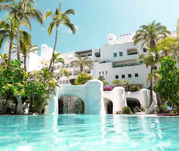 Hvid bygning med palmer omkring og pool i forgrunden. (AI-genereret tekst)