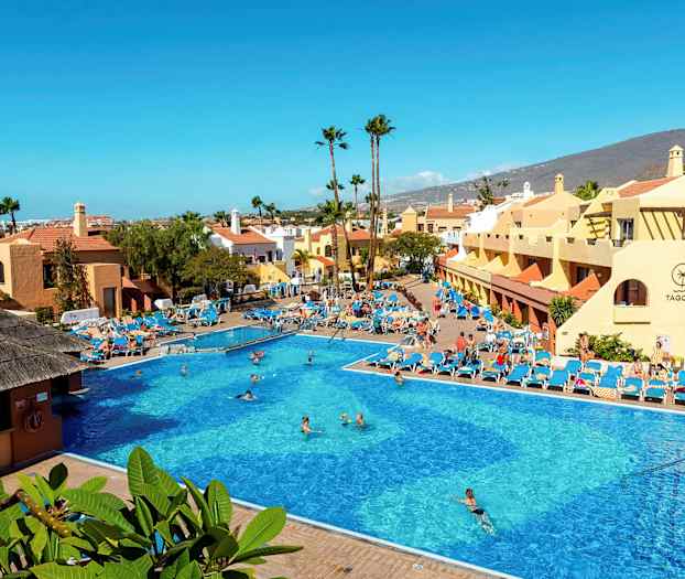 Stor udendørs pool omgivet af solsenge og bygninger med bjerge i baggrunden. (AI-genereret tekst)