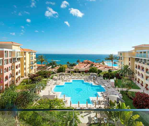 Billede af et hotelområde med en pool omgivet af liggestole og parasoller. Hotelbygningerne er placeret på begge sider, og i baggrunden ses havet. (AI-genereret tekst)